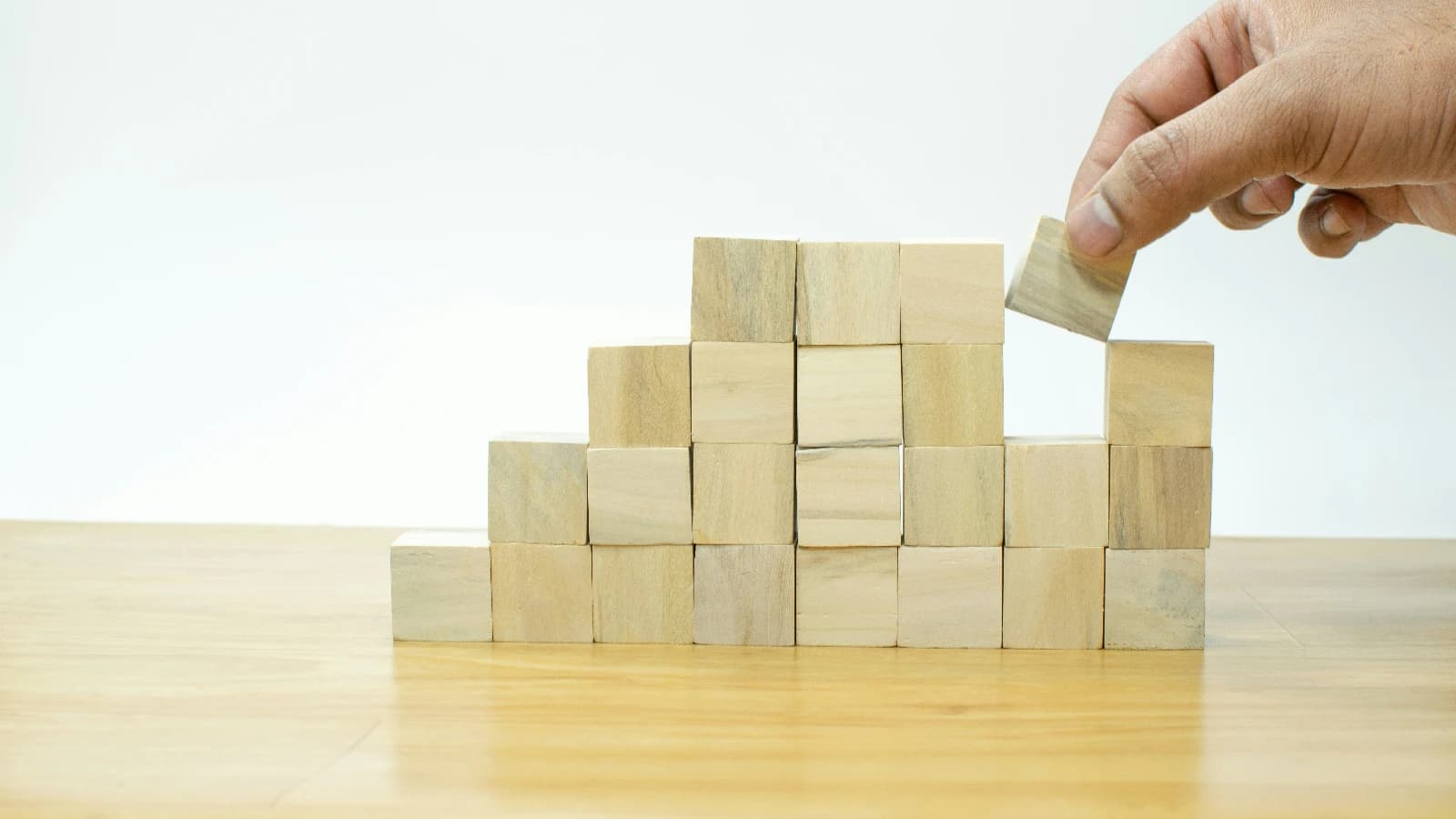 Hand placing a wooden cube on top of a stepped wall of building blocks.