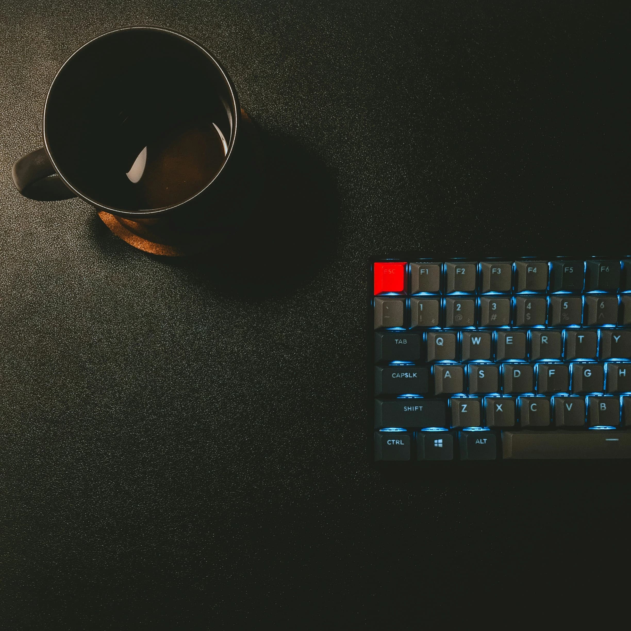 A cup of black coffee and a blue-backlit mechanical keyboard on a dark desk.