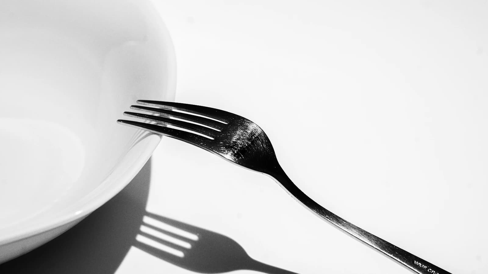 Black-and-white close-up of a metal fork beside a plate, casting a long shadow.