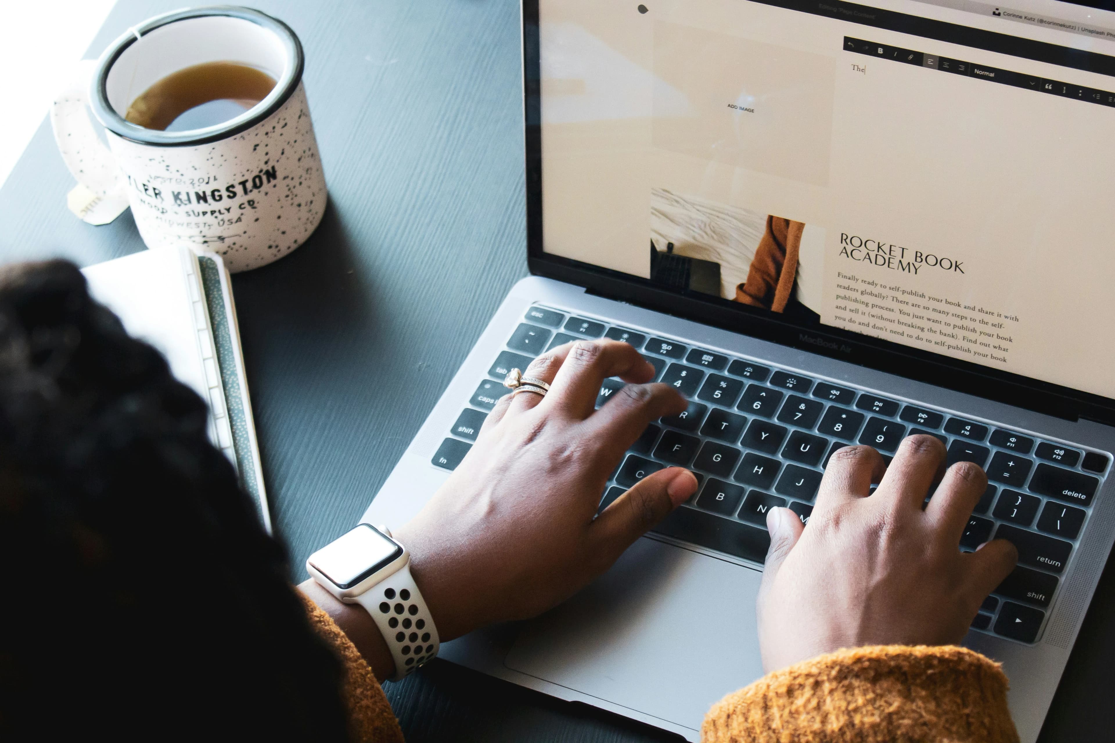 Hands typing on a laptop beside a notebook and a mug of tea.